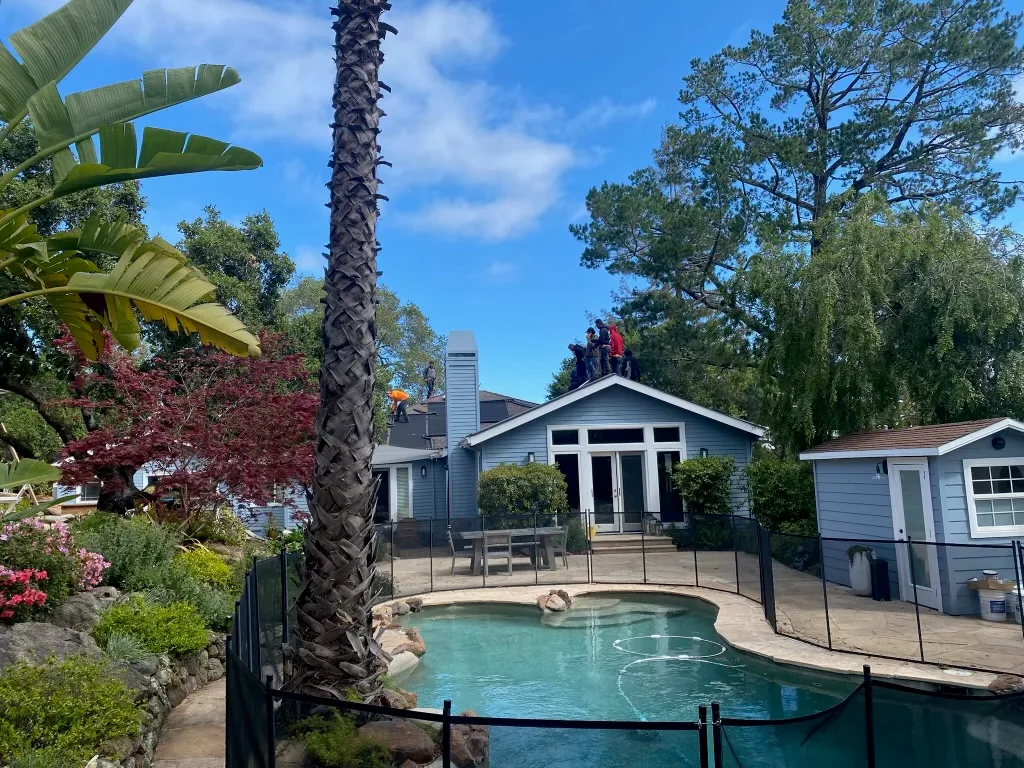 Crew replacing the roof on a gray lap-siding home overlooking a pool and poolside cabana
