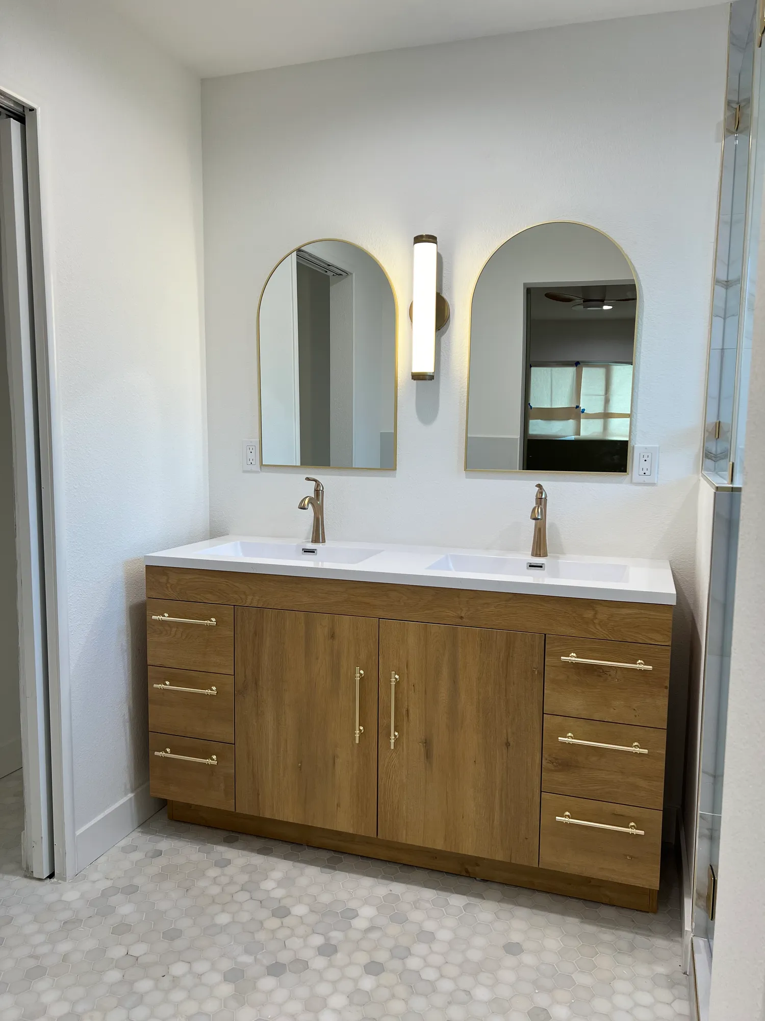 Bathroom double vanity in white oak with arched mirrors, brushed gold faucets, and marble hex floor tile
