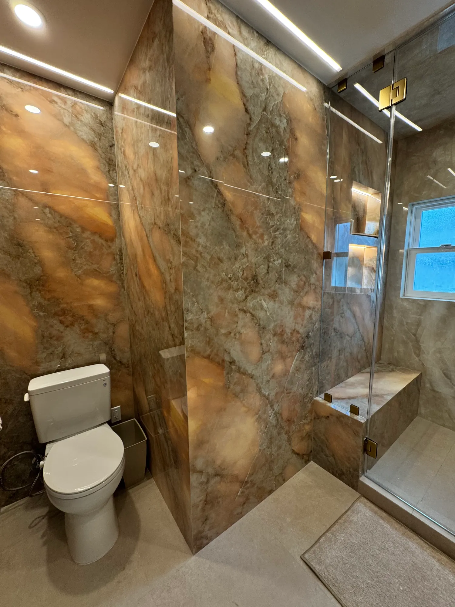 Primary bathroom finished in book-matched quartzite slab with a glass shower enclosure and toilet alcove