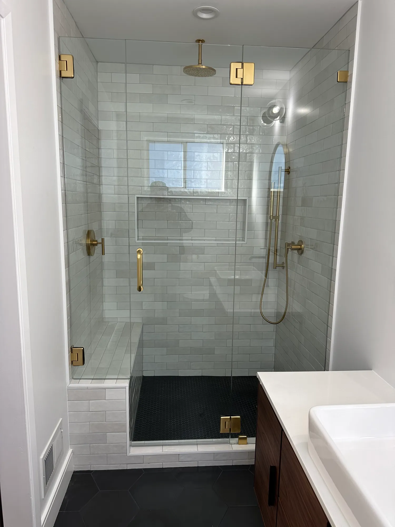 Walk-in glass shower with marble subway tile, brushed gold fixtures, a tiled bench, and black hex floor tile